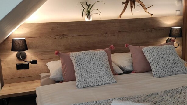 A cozy bedroom with a sloped ceiling, a wooden headboard, a large bed with multiple pillows, a bedside table with a lamp, and a decorative tree trunk on the wall.