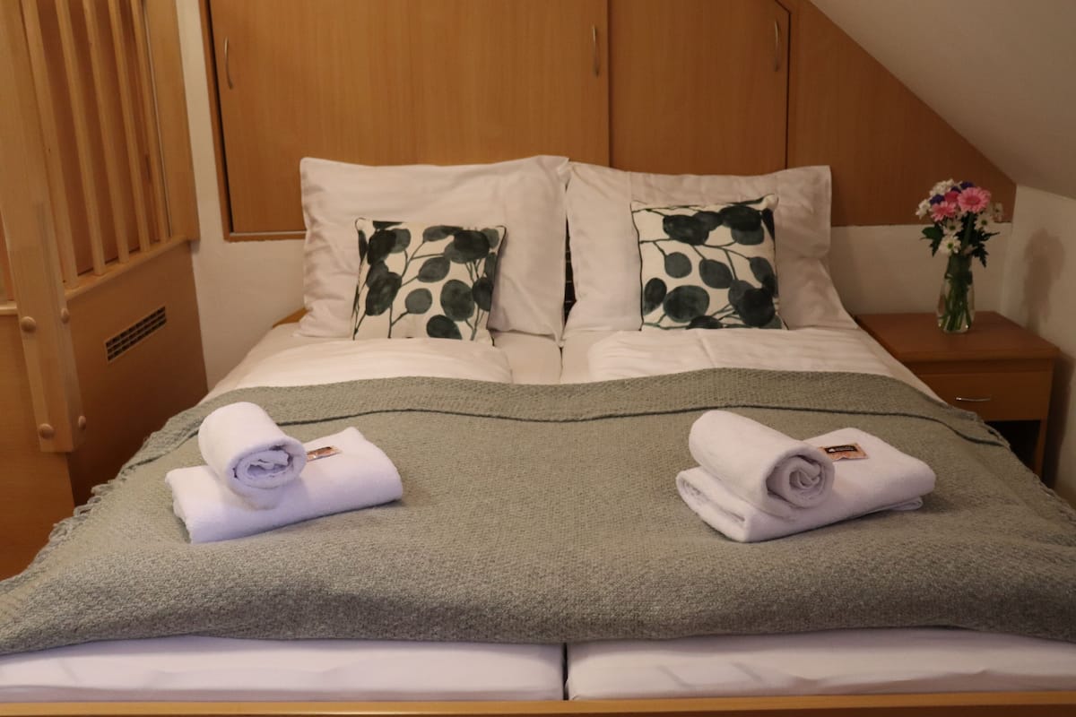 Un lit bien fait, recouvert d’une couverture verte, avec deux oreillers à motifs de feuilles et deux serviettes enroulées posées sur le lit.