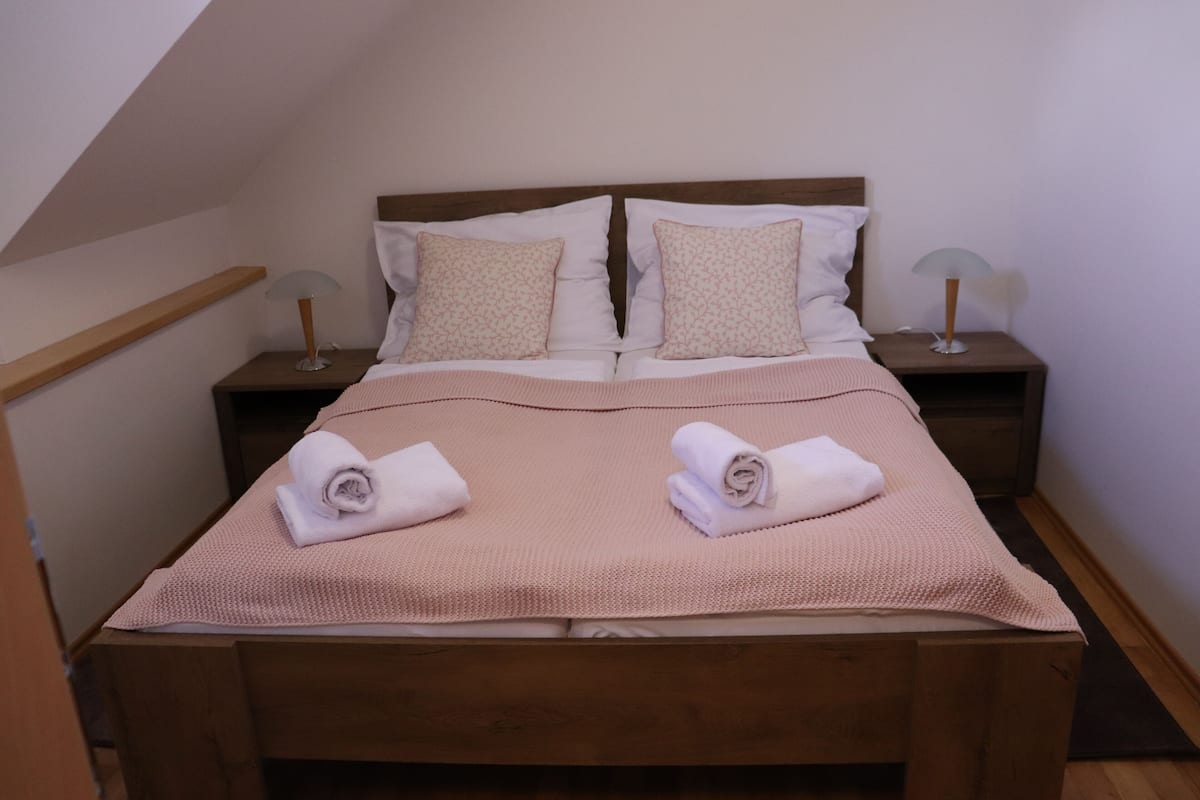 Un lit bien fait, recouvert d’une couverture rose et agrémenté de deux serviettes enroulées, une tête de lit en bois, deux tables de chevet avec des lampes et un banc en bois au pied du lit.