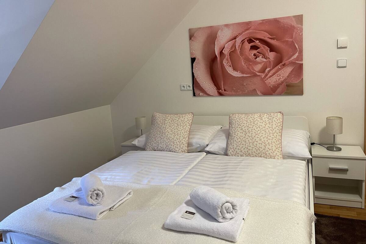 Un lit bien fait, avec du linge de lit blanc et deux serviettes enroulées, un tableau représentant une rose accroché au mur, et une table de chevet avec une lampe.