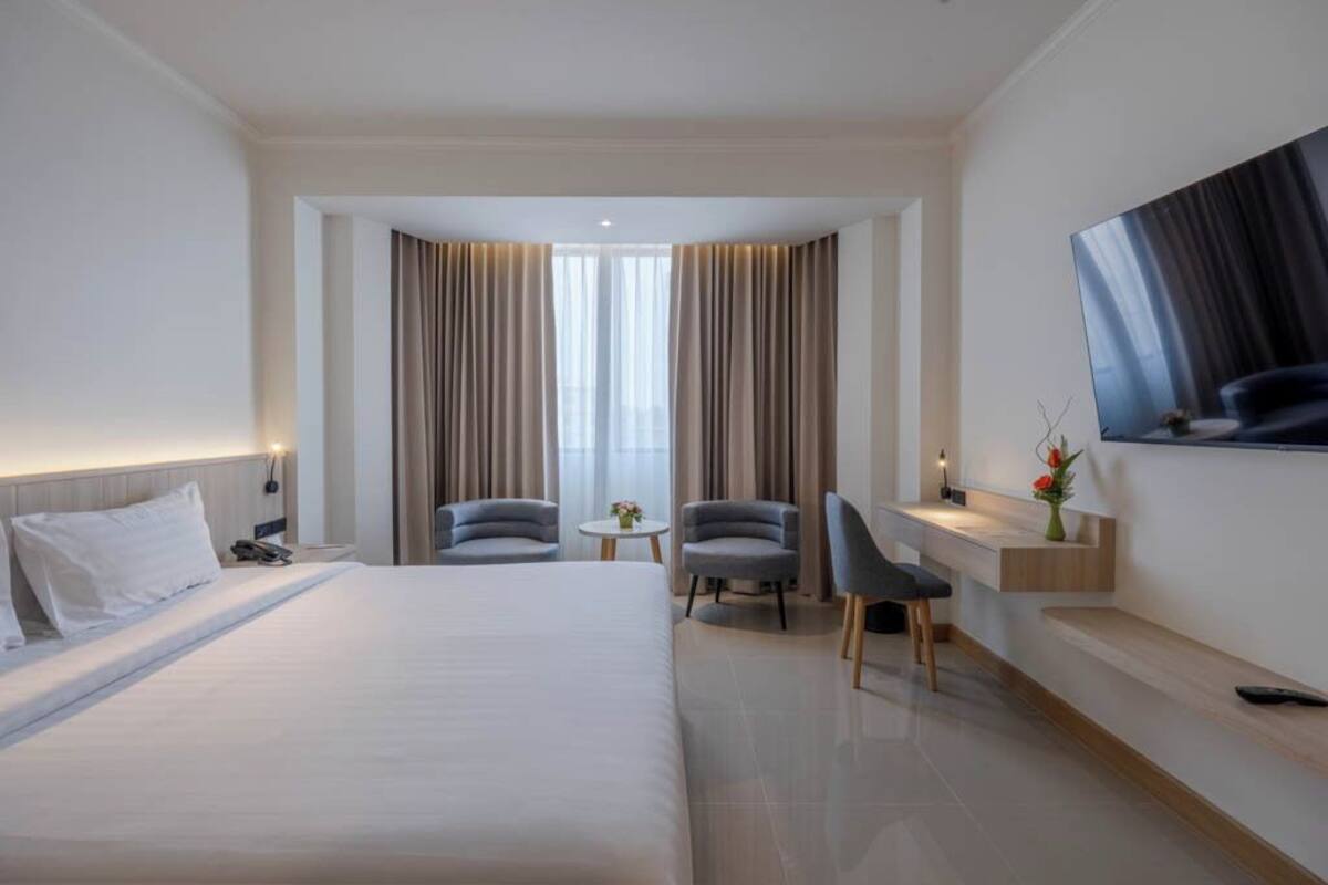 A hotel room with a large bed, two armchairs, a desk, a TV, and a window with curtains.