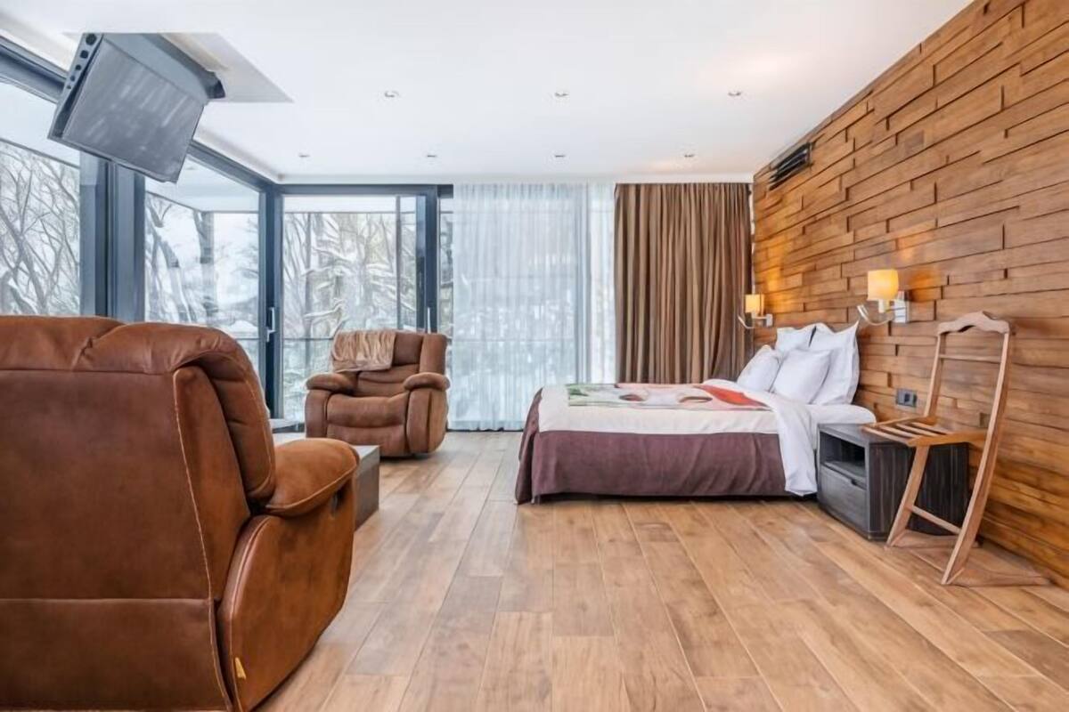 A modern hotel room with a large bed, a leather armchair, a wooden wall, and a large window.