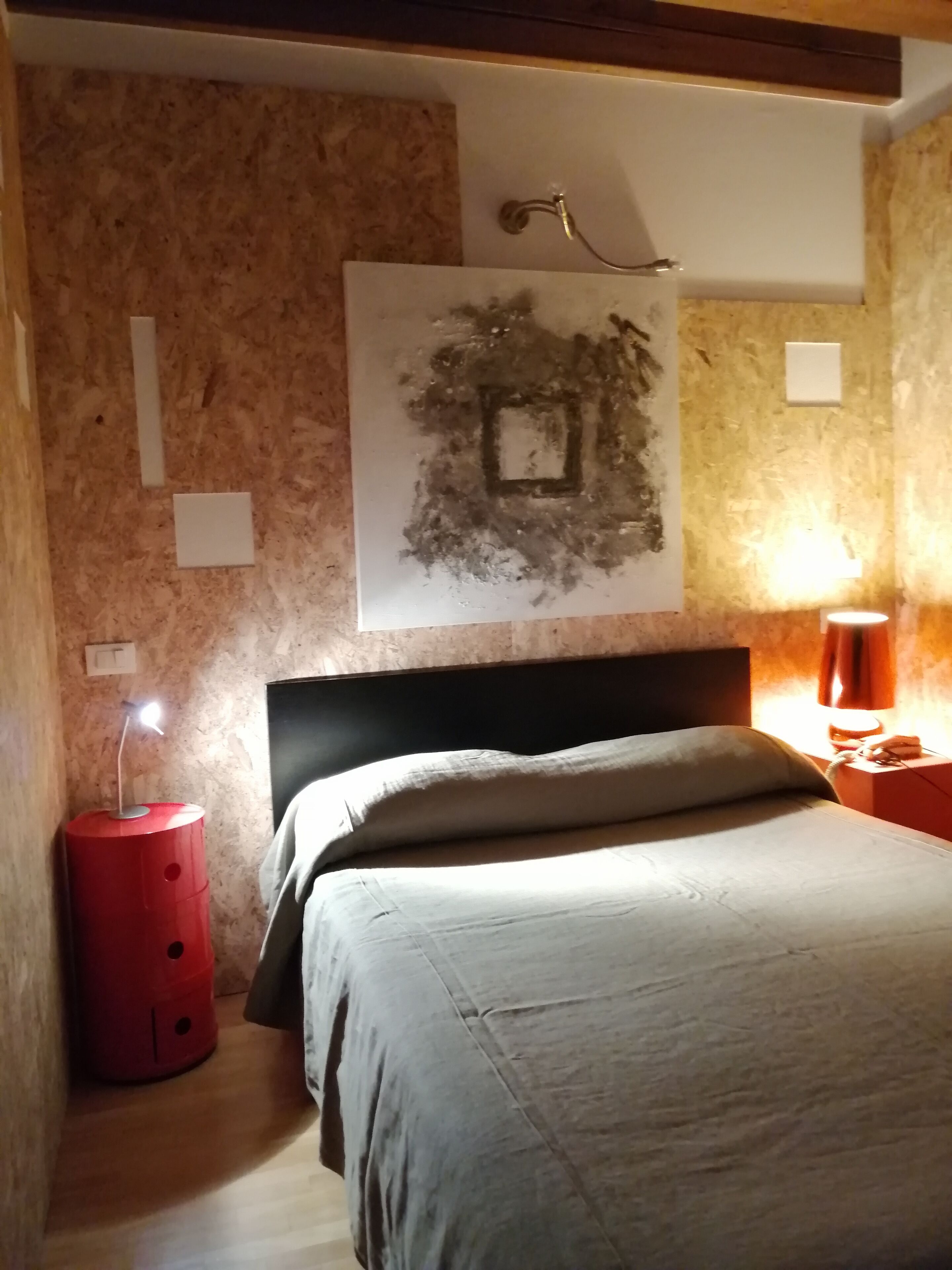 Een slaapkamer met een bed, een rode bijzettafel, een wandlamp en een kunstwerk aan de muur.