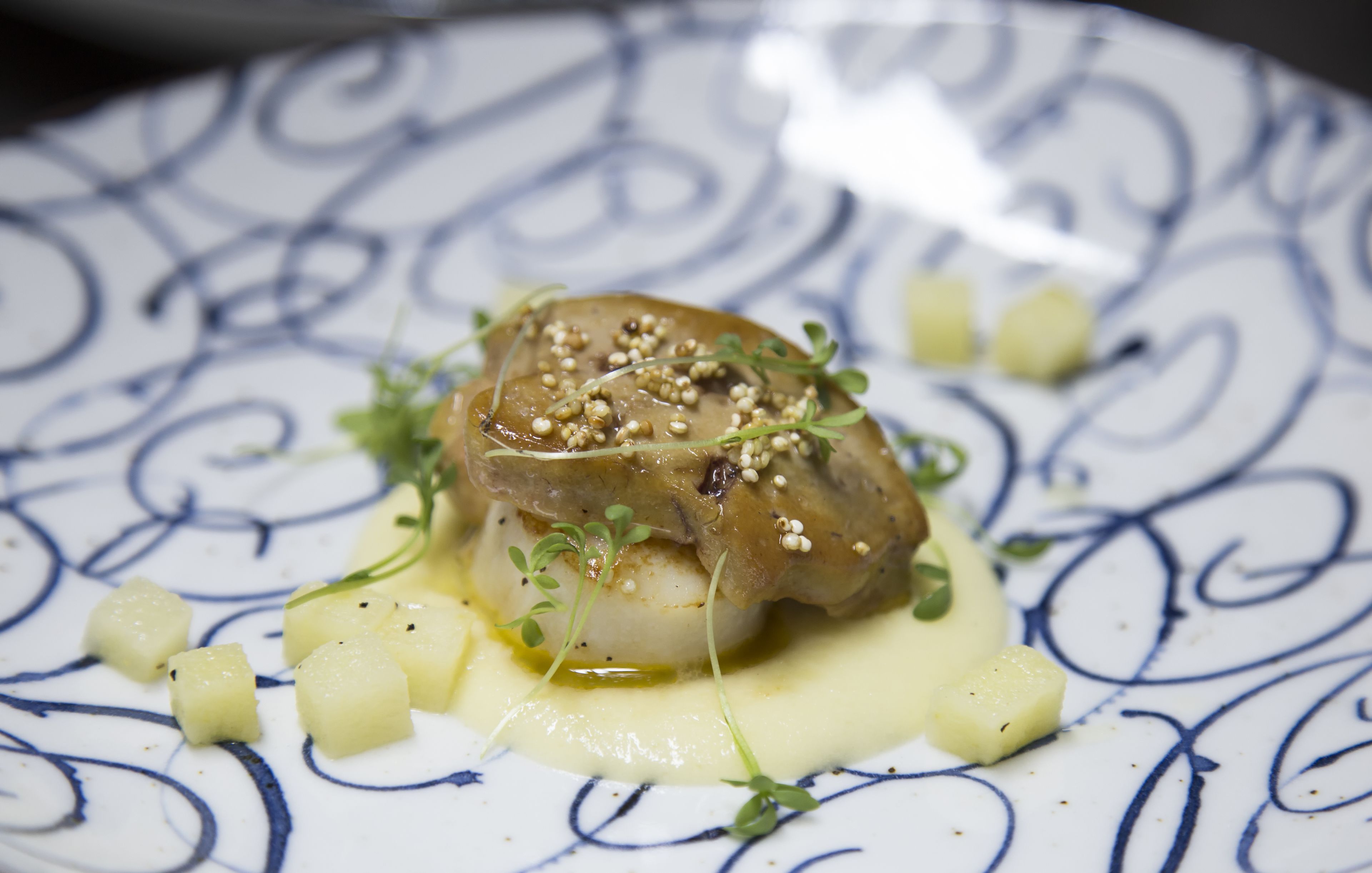 Un plato con un vieiras sellada, sobre un plato estampado, adornado con brotes tiernos y semillas de sésamo, acompañado de patatas en cubos y salsa cremosa.