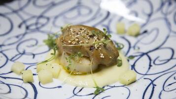 Un plato con un vieiras sellada, sobre un plato estampado, adornado con brotes tiernos y semillas de sésamo, acompañado de patatas en cubos y salsa cremosa.
