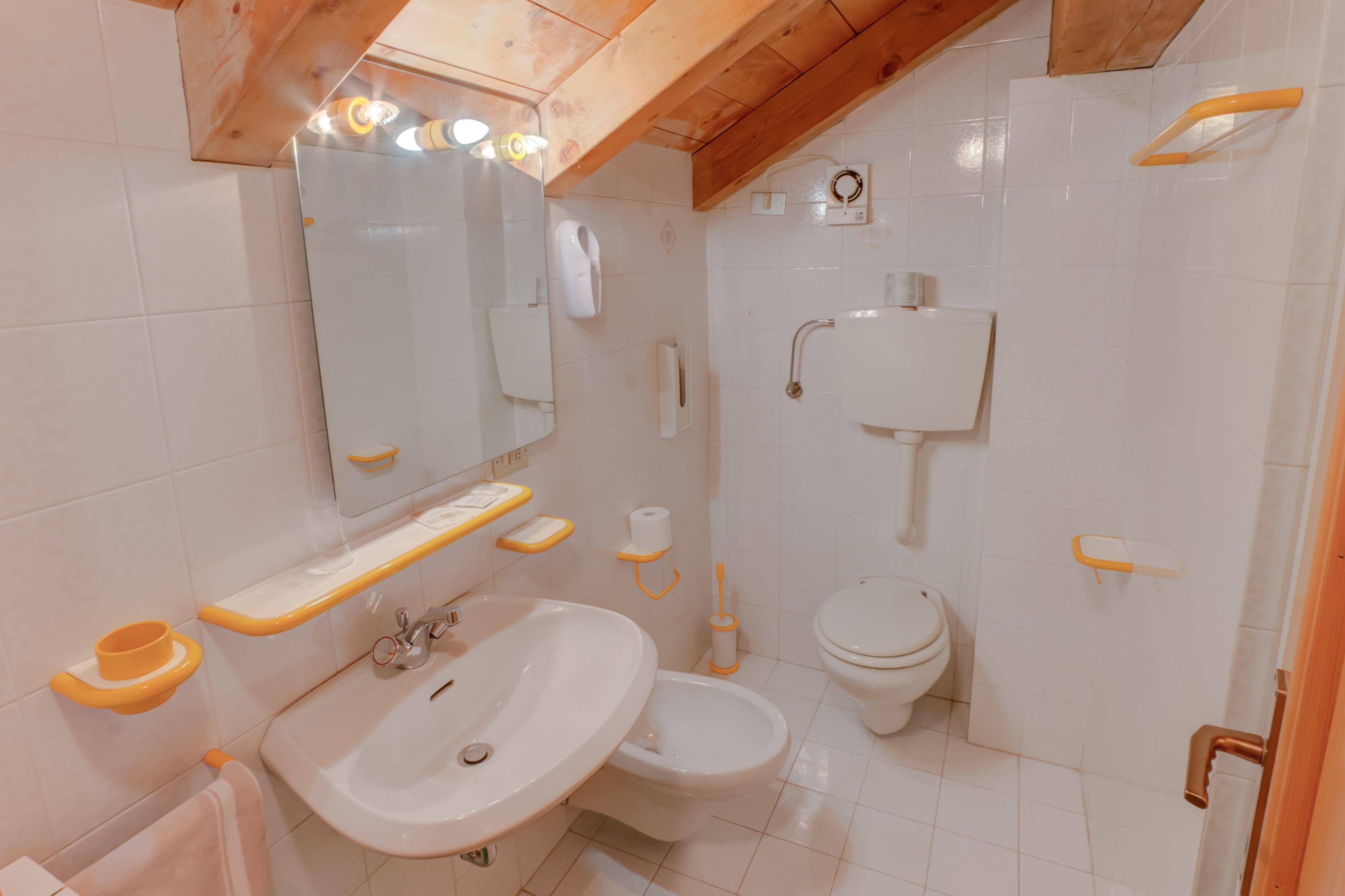 A bathroom with a sink, toilet, and bidet. It has white tiles and wooden beams on the ceiling.
