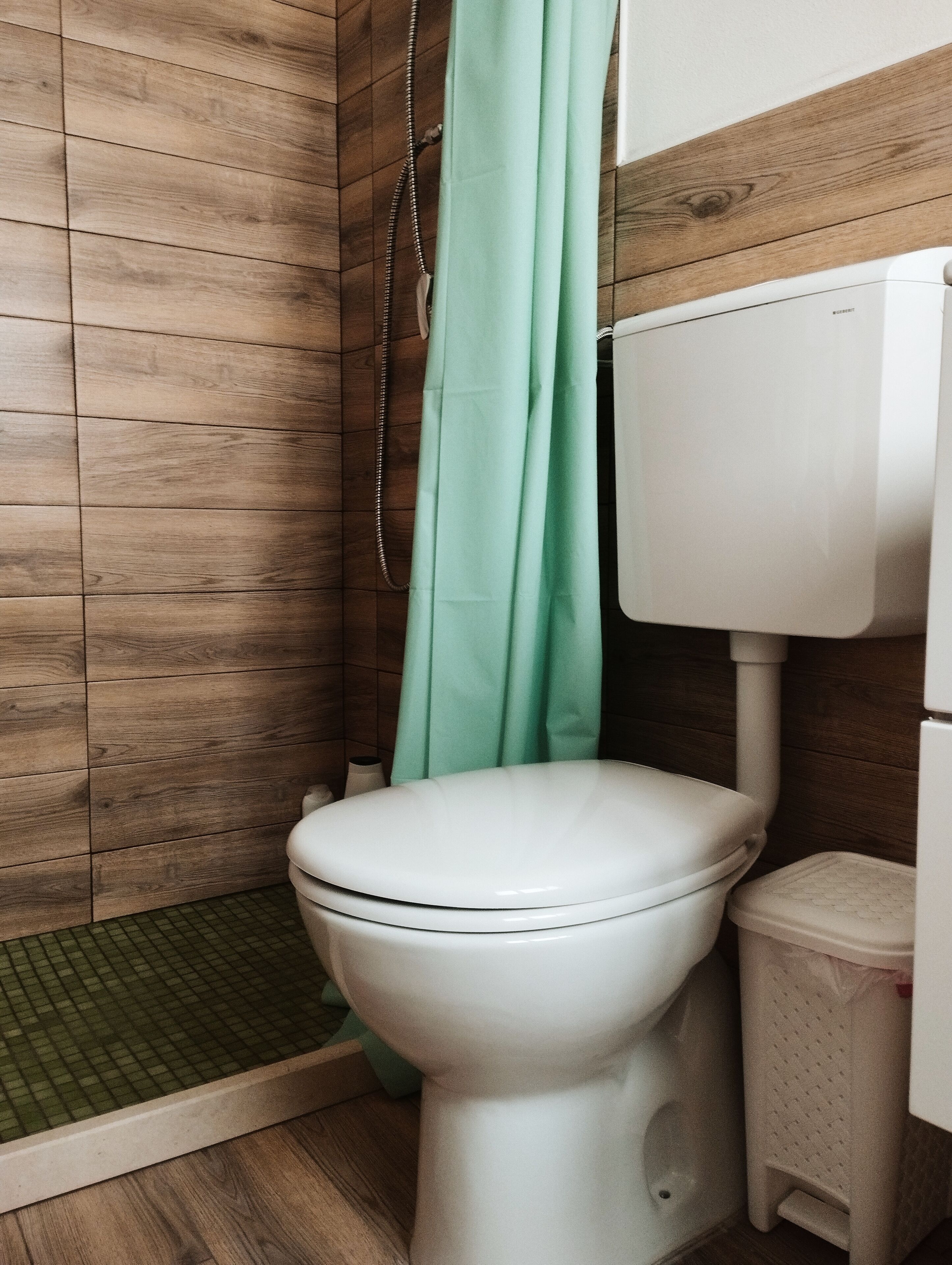 A bathroom with a wooden wall, a white toilet, a green shower curtain, and a white trash bin.