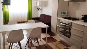 A compact kitchen with a gas stove, oven, and a small countertop. There is a dining table with chairs and a sofa in the adjacent living area. - Thomas'home (Comiso)