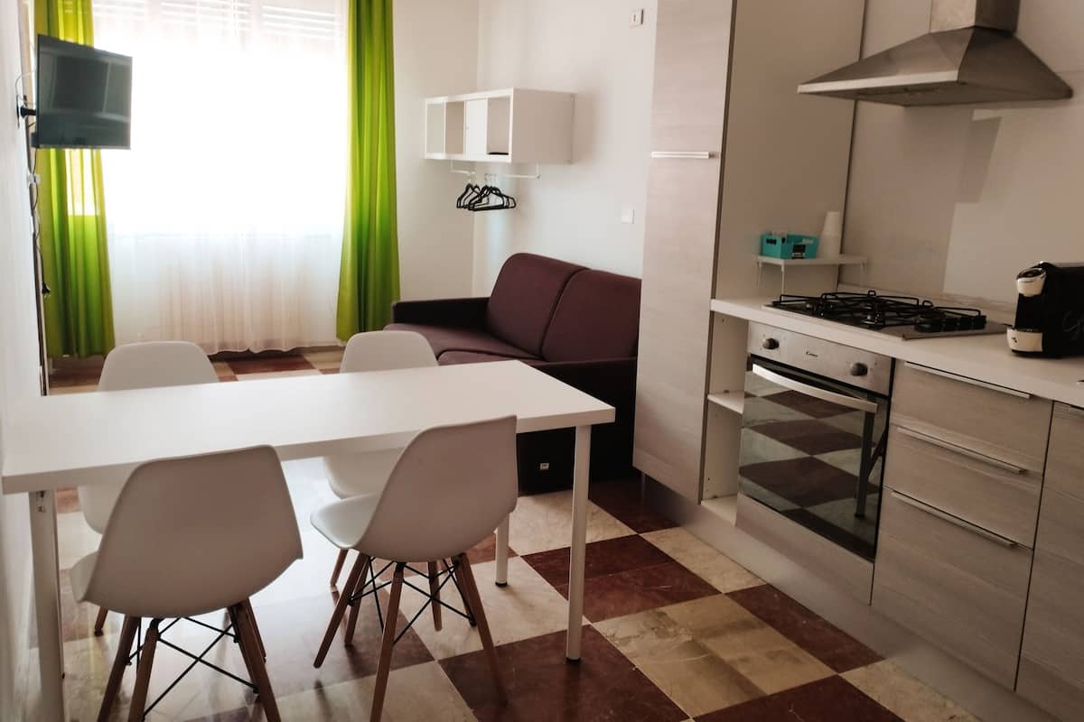 A compact kitchen with a gas stove, oven, and a small countertop. There is a dining table with chairs and a sofa in the adjacent living area.