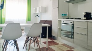 A modern kitchen with white cabinets, a stove, and a sink. There is a dining table with chairs and a window with green curtains. - Thomas'home (Comiso)