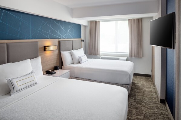 Ein Hotelzimmer mit zwei Betten, einem Fernseher, einem Nachttisch mit Lampe, einem Fenster mit Vorhängen und einem an der Wand montierten Fernseher.