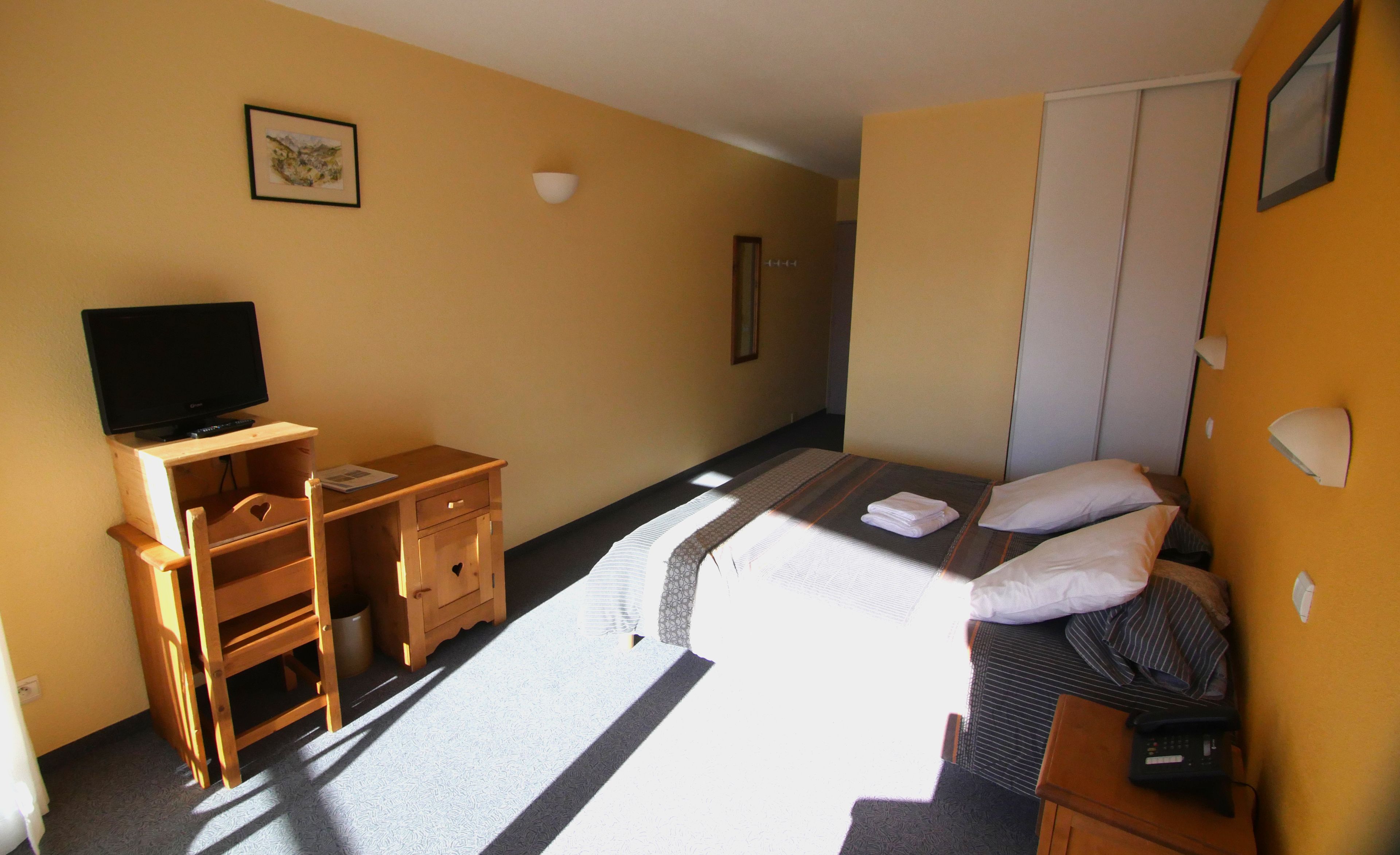 Quarto de hotel com cama, escrivaninha com computador, cadeira e televisão.