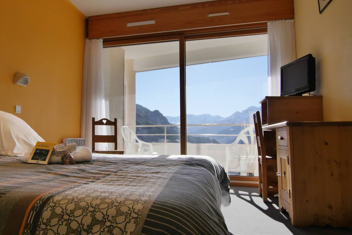 A hotel room with a bed, a television, a wooden dresser, and a balcony with a mountain view.