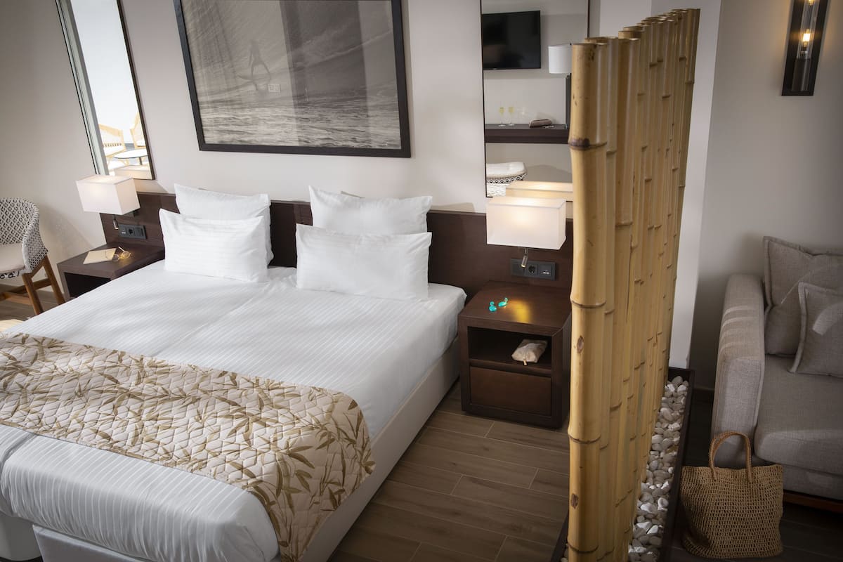 A hotel room with a large bed, a bedside table with a lamp, a small table with a bottle, a chair, a sofa, and a decorative bamboo screen.