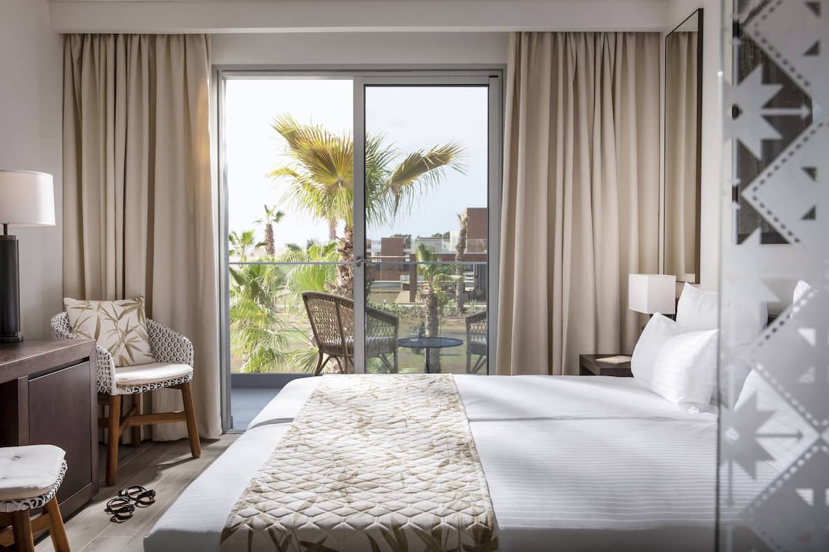 A hotel room with a bed, a desk, a chair, and a balcony with a view of palm trees and buildings.