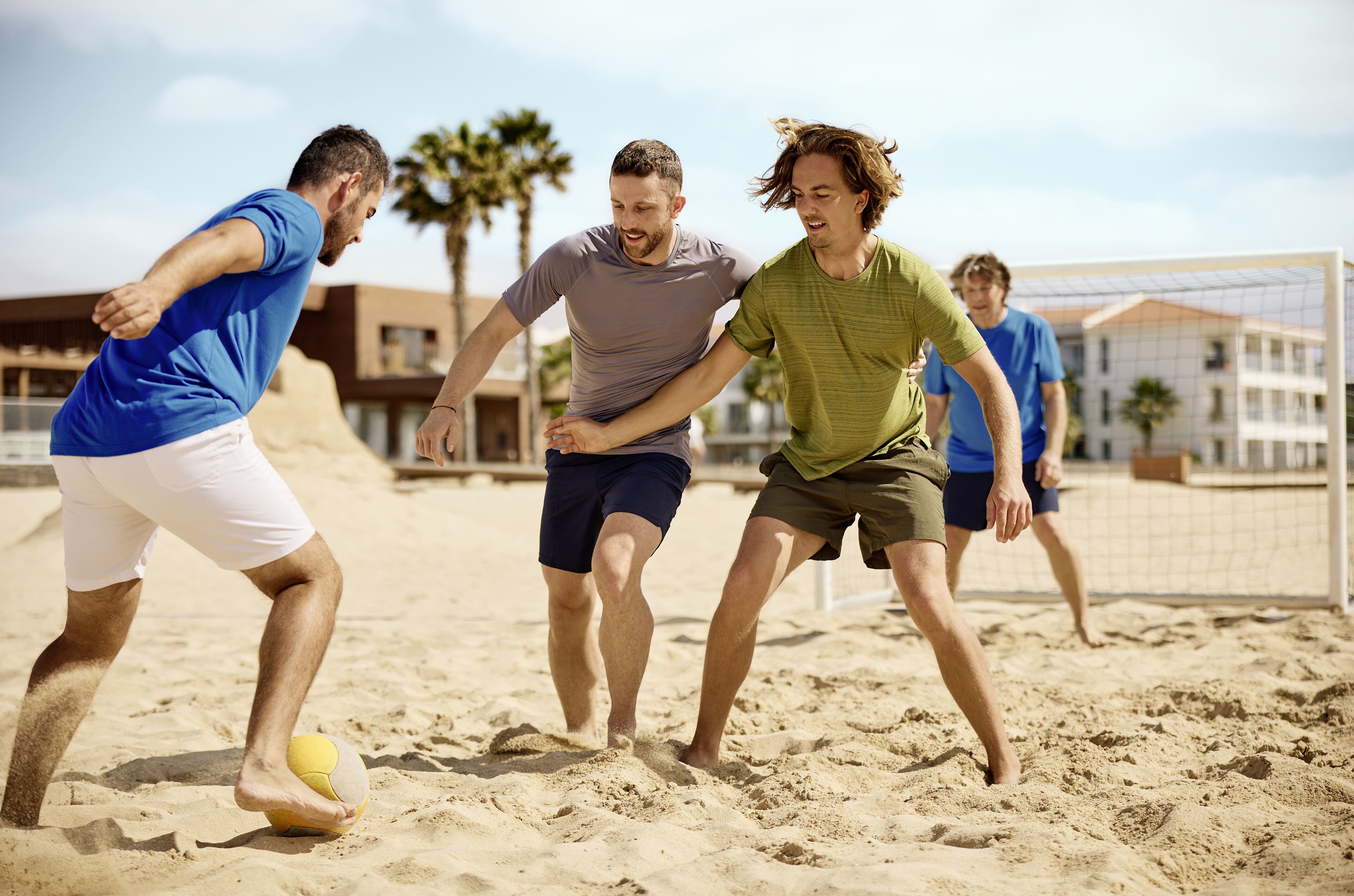 Un grupo de personas juega voleibol de playa en una cancha arenosa, con edificios al fondo.