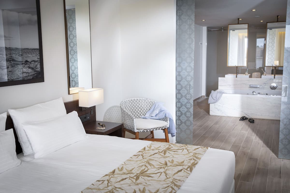 A hotel room with a large bed, a chair, a desk, a lamp, and a bathroom with a mirror and sink.