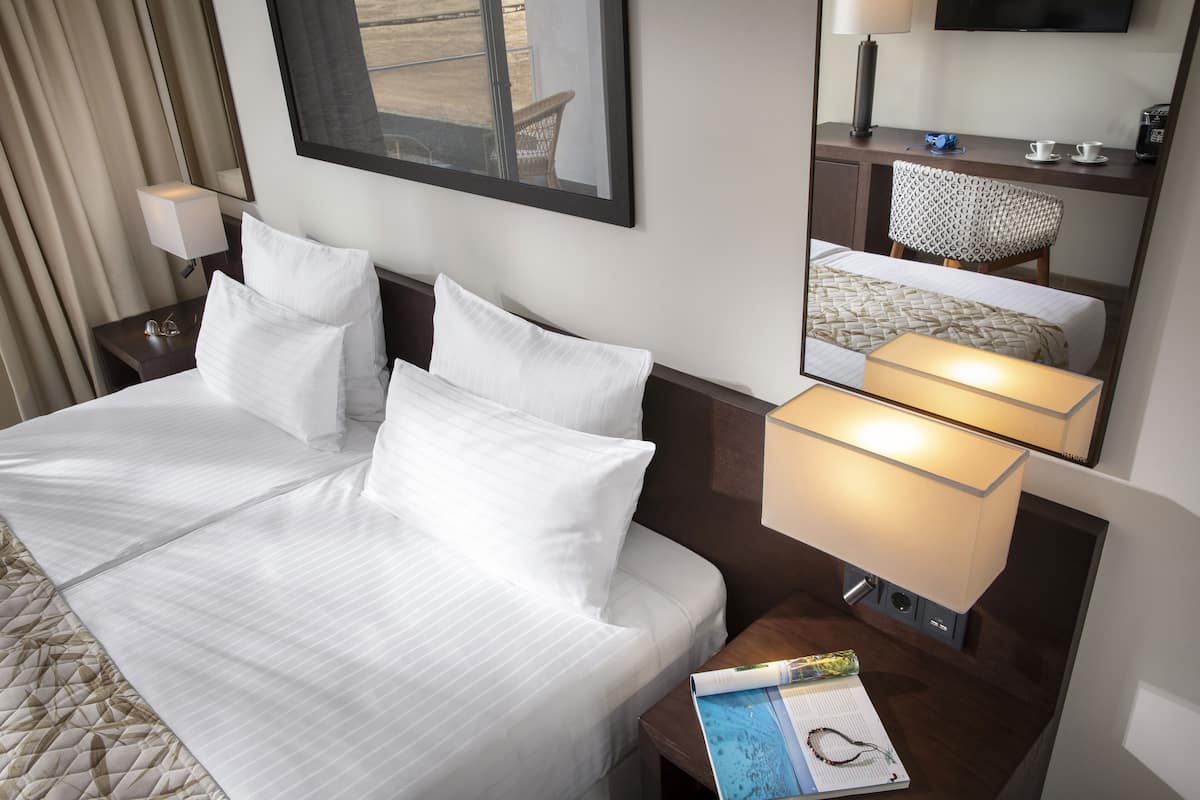 A hotel room with a neatly made bed, a bedside table with a lamp, a chair, a desk with a computer, and a large mirror.