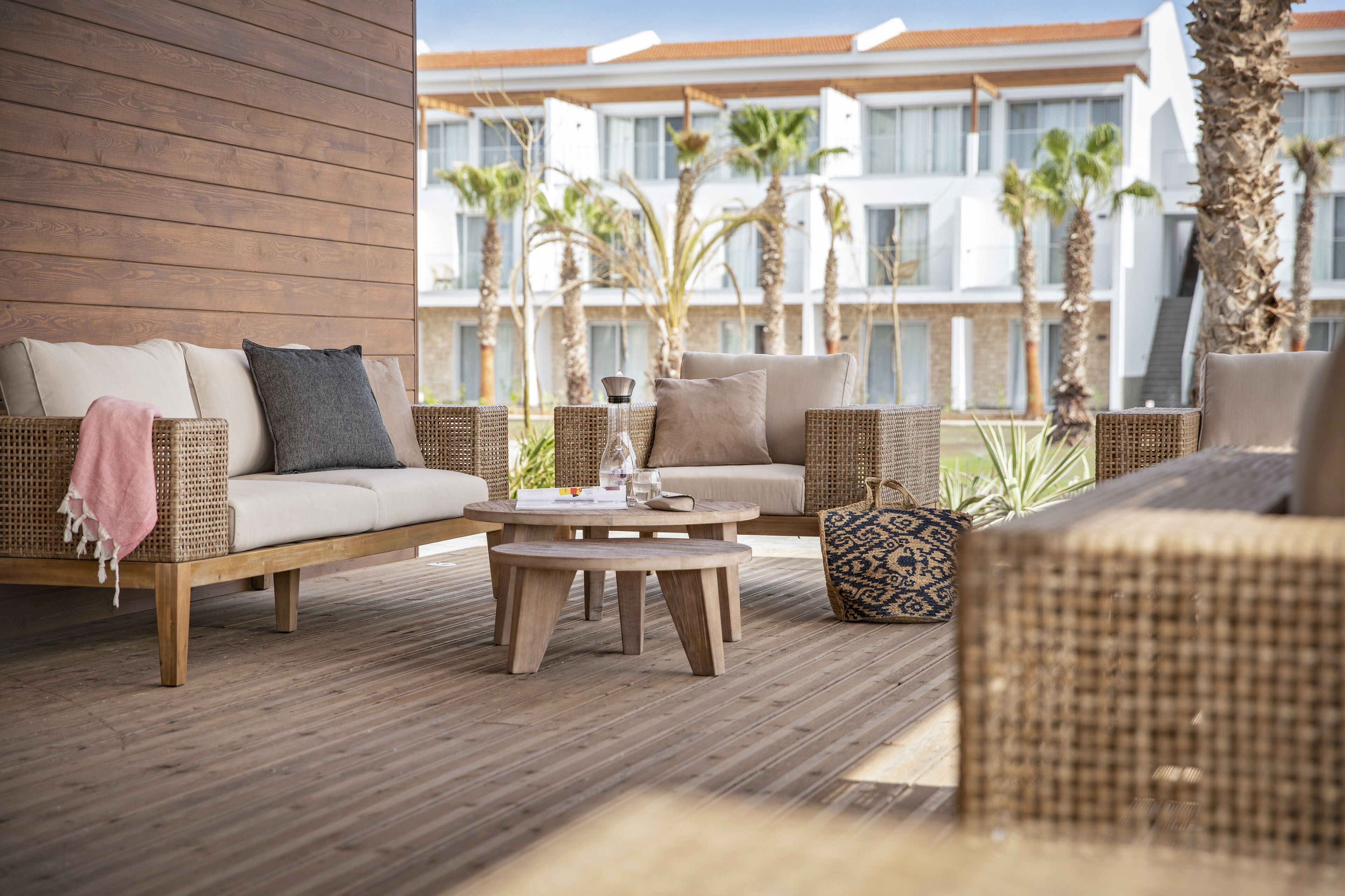 Una terraza de madera con muebles de mimbre, incluyendo un sofá y sillas, y una mesa redonda con superficie de vidrio.