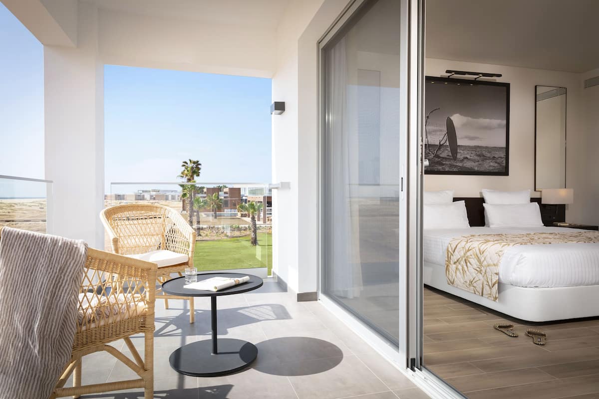 A balcony with wicker chairs and a table, a bedroom with a bed and a large window, and a view of the beach and buildings.