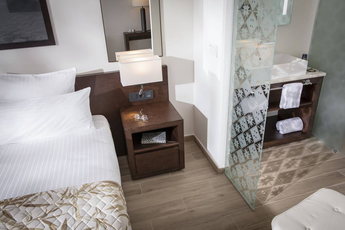 A hotel room with a bed, a nightstand with a lamp, a bathroom with a mirror, and a shower area.
