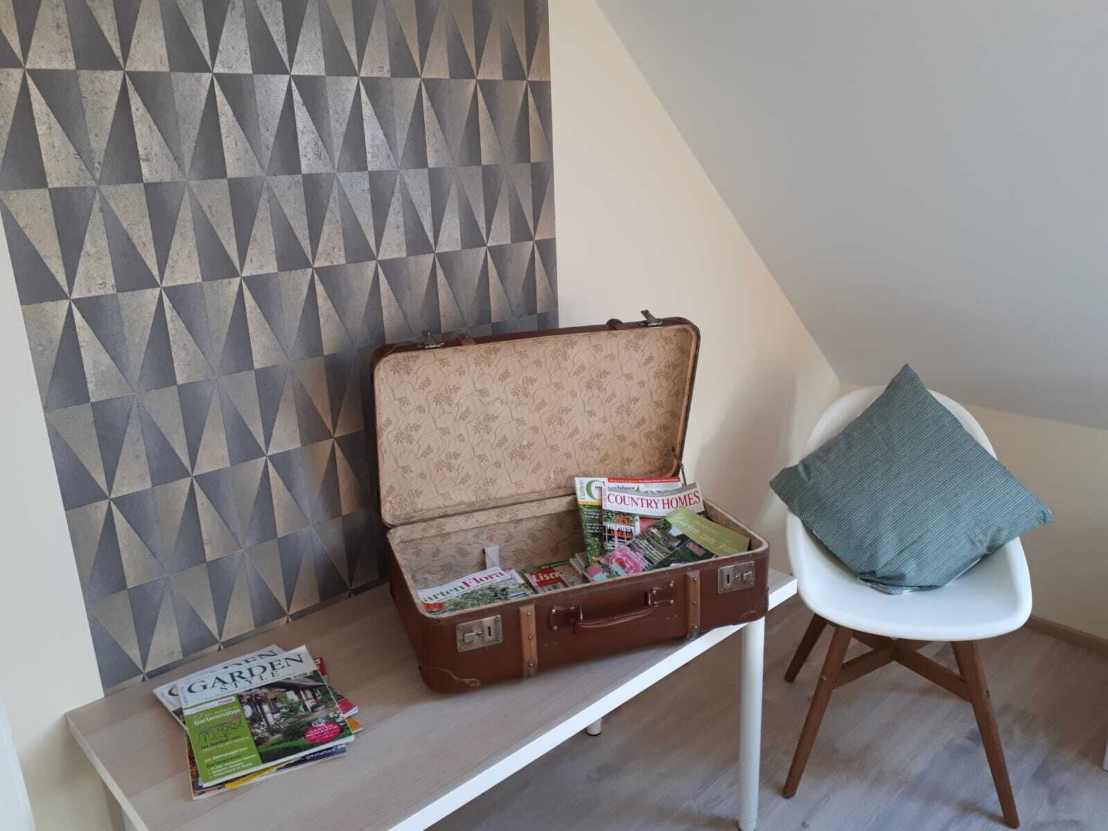 A room with a geometric-patterned wall, a wooden chair with a cushion, and a suitcase filled with magazines on a white table.
