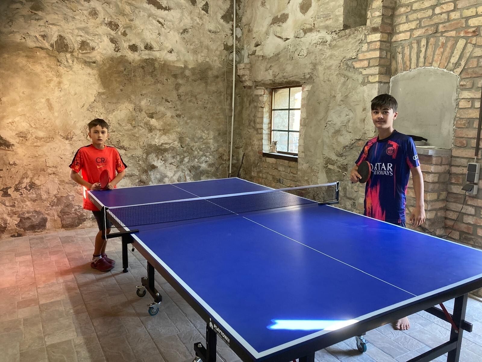 Zwei Personen spielen Tischtennis in einem Raum mit rustikaler Ziegelwand und Fenster.