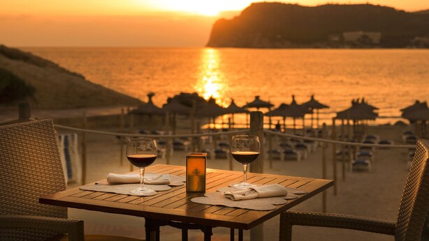 A table with two glasses of red wine, a candle, and a menu on a wooden table overlooking a beach with umbrellas and lounge chairs. - Hapimag Resort Paguera (Calvia)