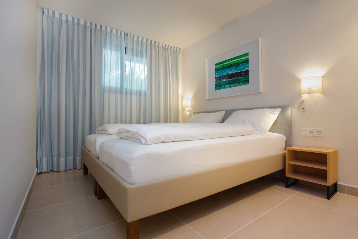 A neatly made bed in a hotel room with a view of the outdoors through sheer curtains, a bedside table with a lamp, and a painting on the wall.