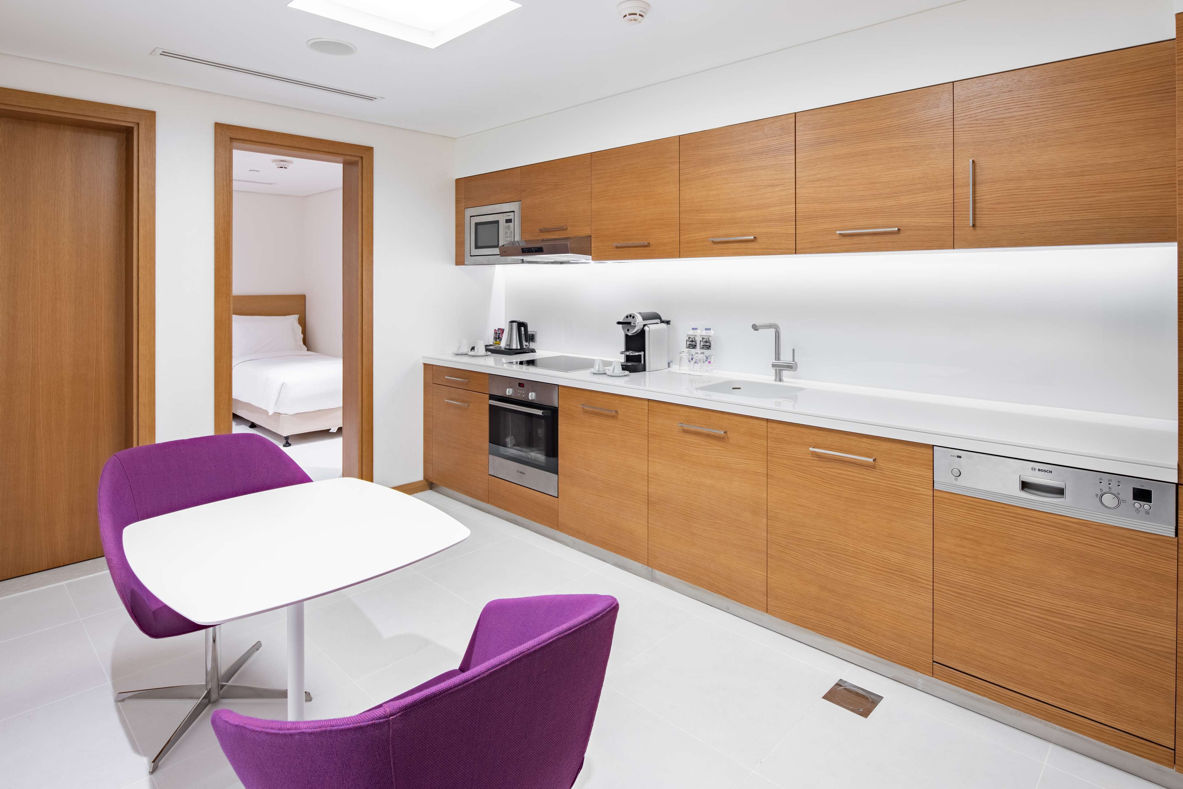 Una cocina moderna con armarios de madera, una encimera blanca y un lavavajillas Bosch. Se ve una silla morada y una cama en la habitación contigua.