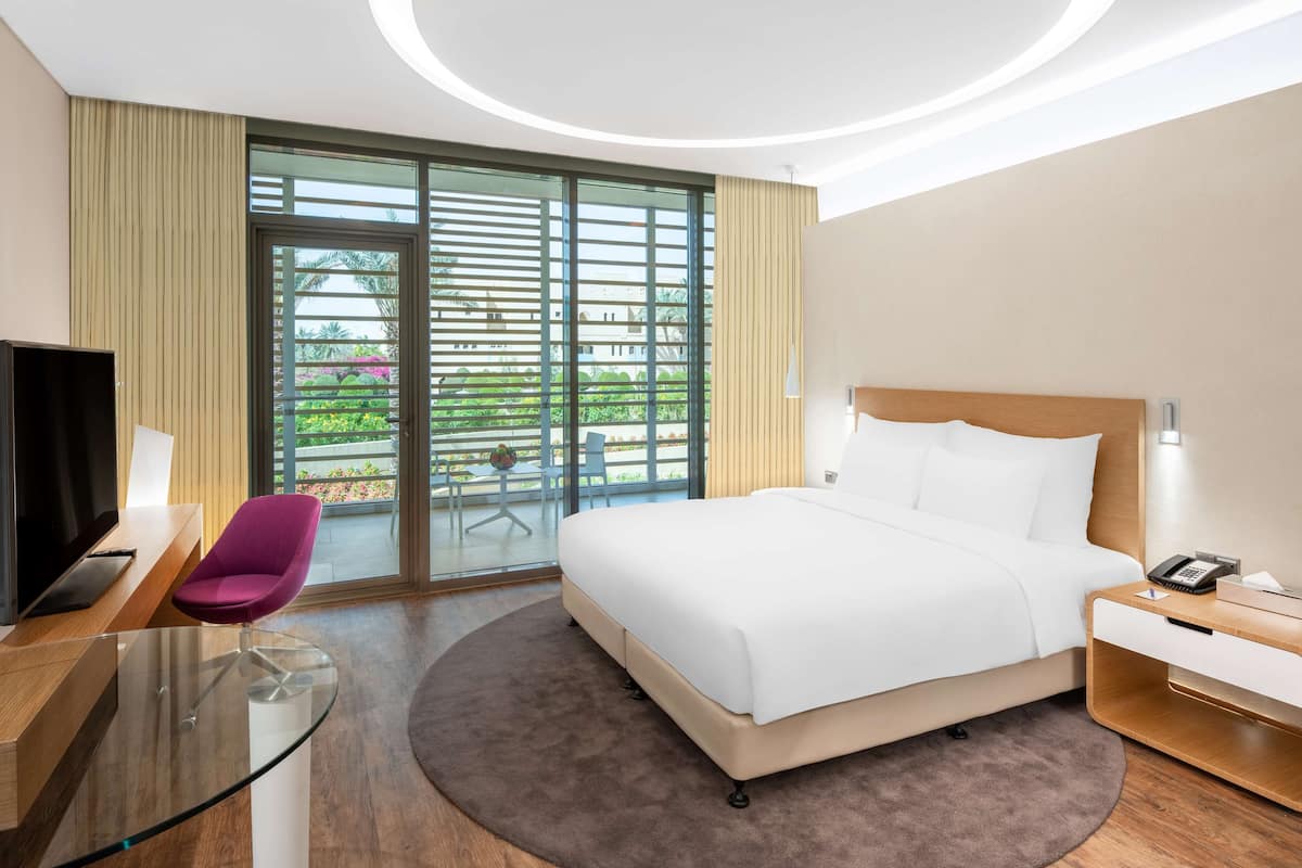 Habitación de hotel moderna con una cama grande, un escritorio de vidrio, una silla morada y vistas al exterior a través de una puerta corredera.