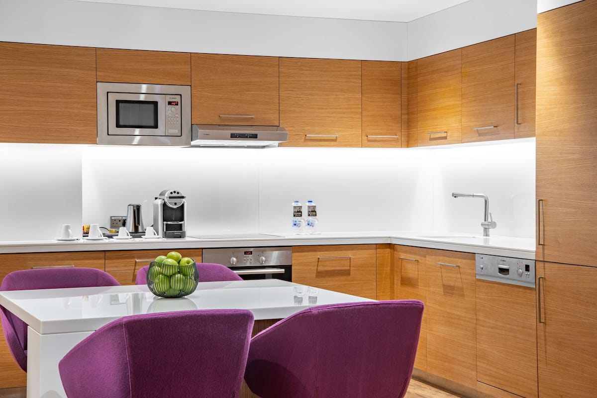 Una cocina moderna con armarios de madera, un microondas de acero inoxidable, un horno empotrado y una encimera blanca. Hay sillas moradas alrededor de una mesa blanca con un cuenco verde.