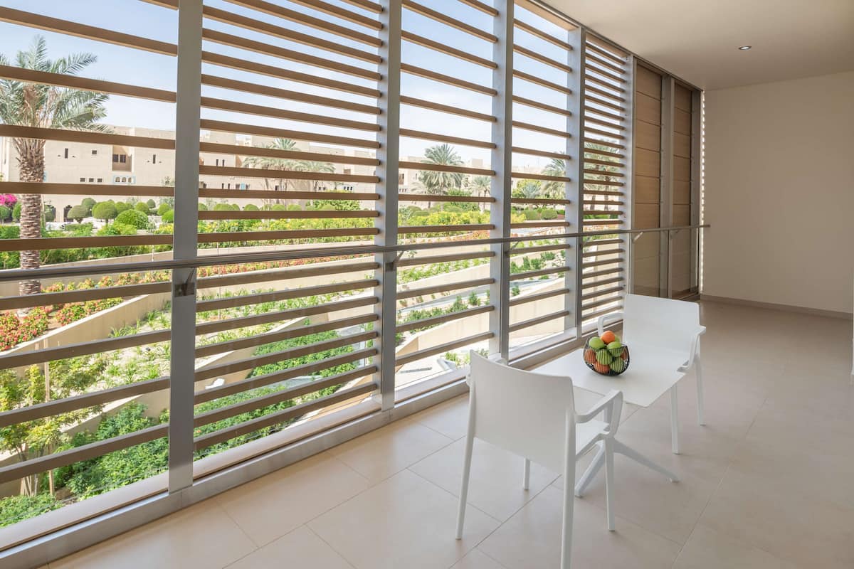 Un balcón con vistas a un jardín y edificios, una mesa blanca con sillas y un frutero.
