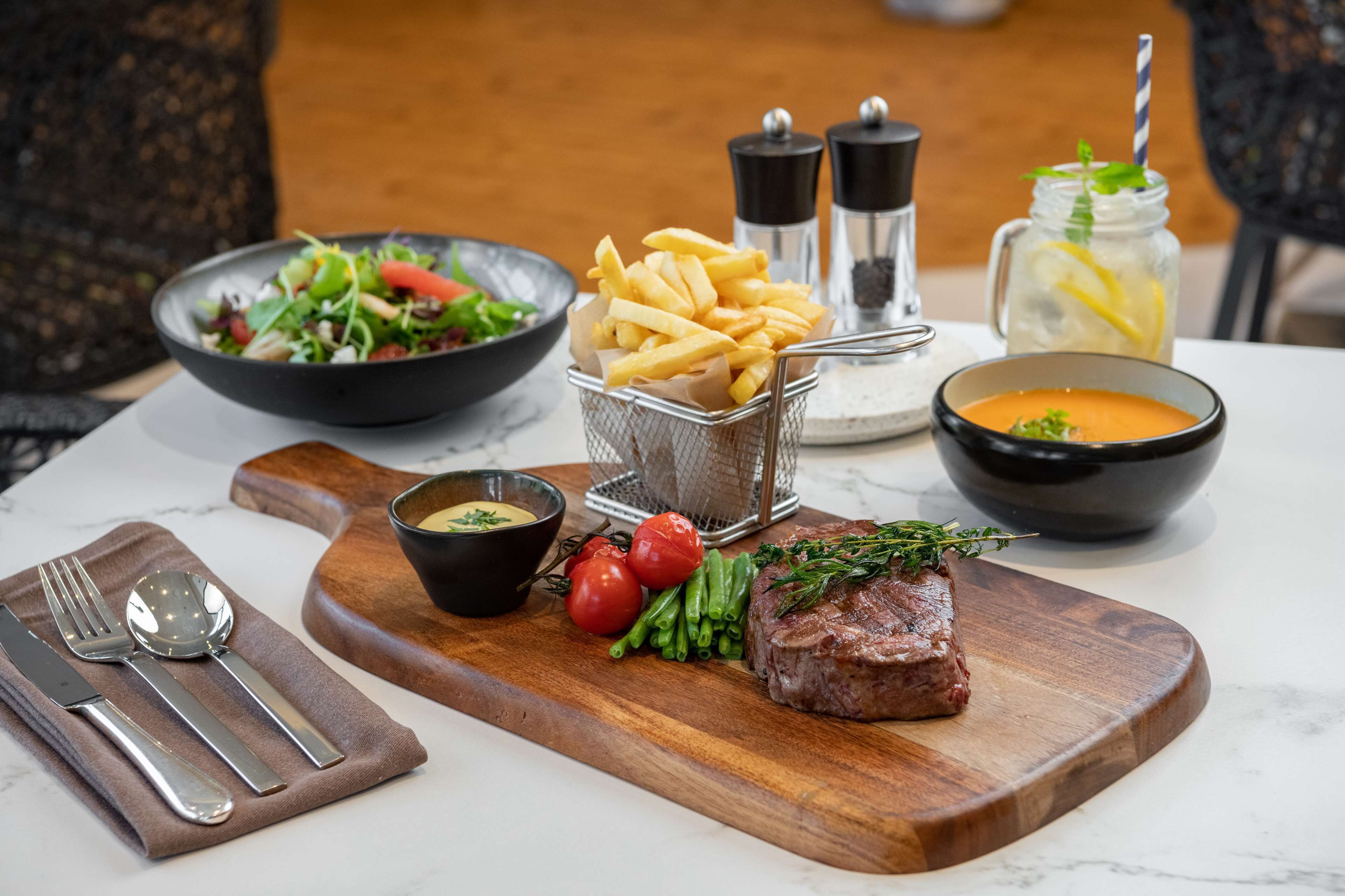 A wooden cutting board with a steak, green beans, tomatoes, and a small bowl of sauce.