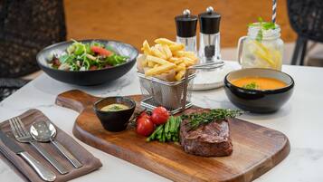 A wooden cutting board with a steak, green beans, tomatoes, and a small bowl of sauce.