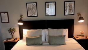 Una cama bien hecha con sábanas blancas, dos almohadas y dos toallas dobladas sobre una colcha verde. Hay obras de arte enmarcadas en la pared sobre la cama y dos lámparas de noche sobre mesitas de madera.