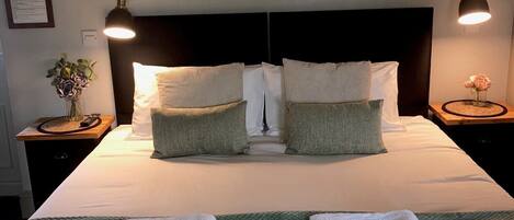 Un lit bien fait, avec des draps blancs, deux oreillers et deux serviettes pliées posées sur un couvre-lit vert. Des œuvres d’art encadrées ornent le mur au-dessus du lit, et deux lampes de chevet sont posées sur des tables de chevet en bois.