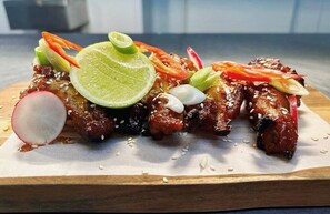 A dish of glazed meat with sesame seeds, garnished with radish, tomato, and green onions, served on a wooden board.
