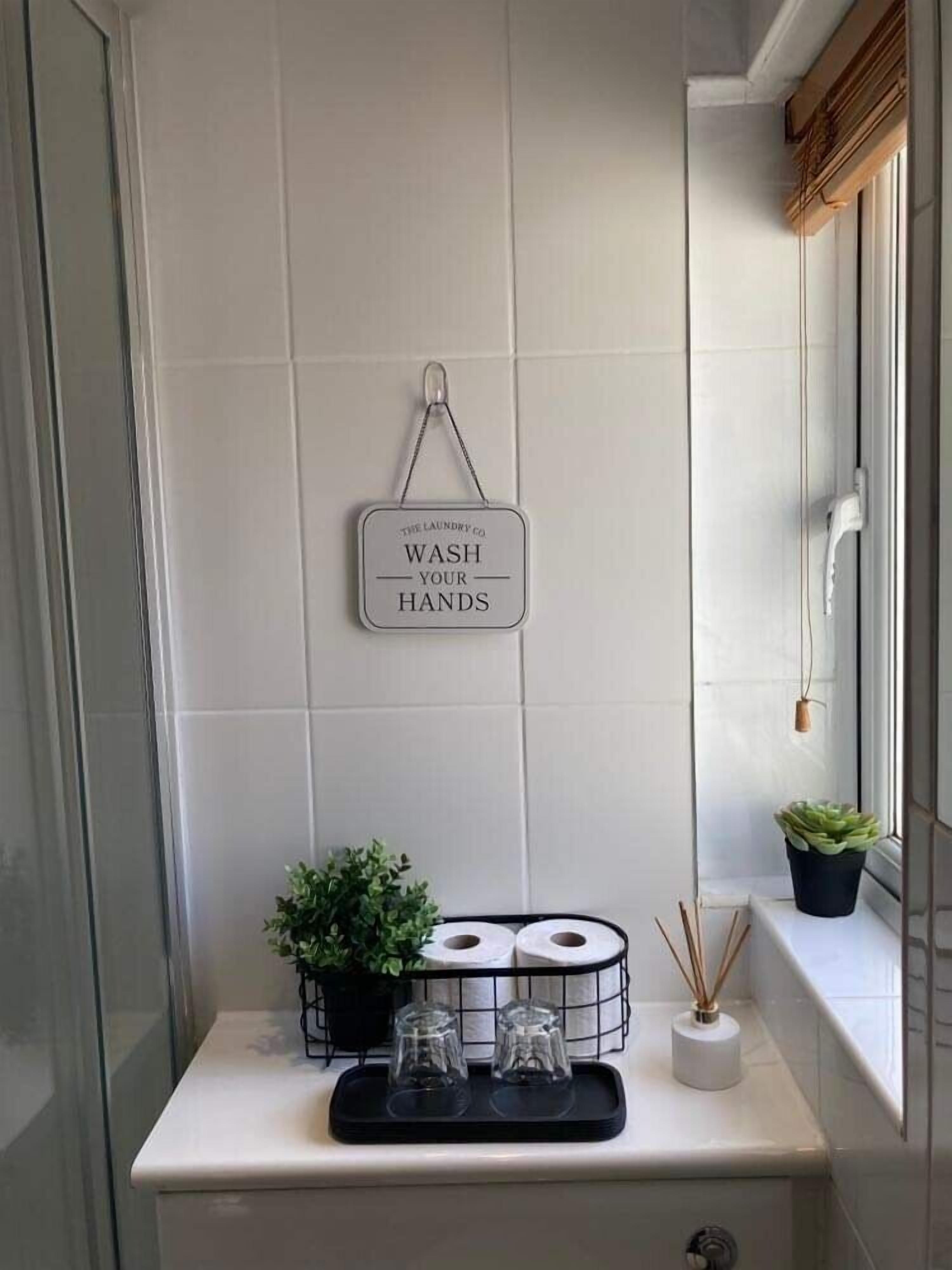 Une salle de bain avec un mur recouvert de carreaux blancs, un panneau indiquant « LAVEZ-VOUS LES MAINS », une petite plante, deux verres et un rouleau de papier toilette posé sur une étagère.