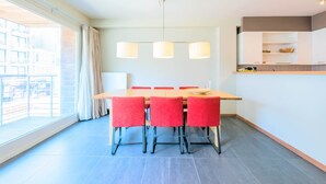A modern dining area with a wooden table and red chairs, a hanging light fixture, and a view of the city through large windows. - Ines 101 (Koksijde)