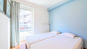 A bedroom with two beds, a window with curtains, and a view of buildings outside. - Ines 101 (Koksijde)