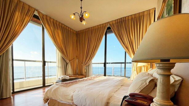 A hotel room with a large bed, a bedside telephone, and a view of the sea through large windows. - Holiday Home Suites (Beirut)