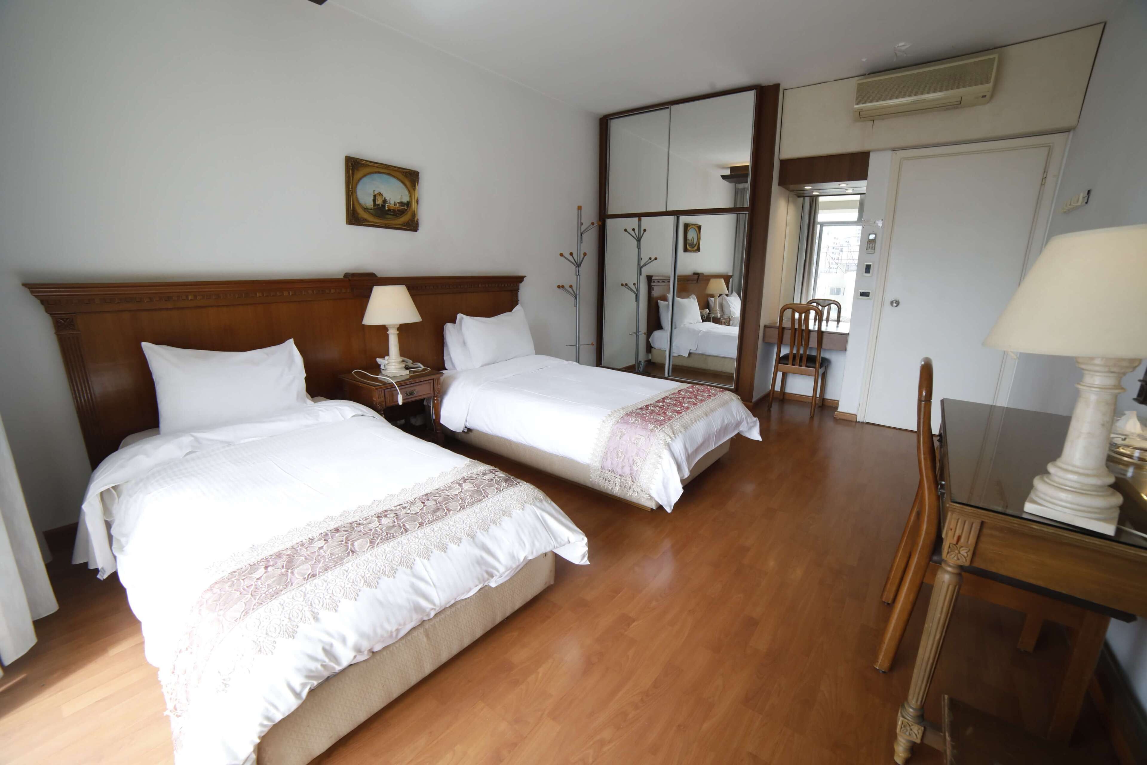 Ein Hotelzimmer mit zwei Betten, einem Holz-Kopfteil, einem Nachttisch mit Lampe, einem Spiegel und einem Esstisch mit Stühlen.