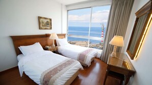 A hotel room with two beds, a nightstand, a desk, and a large window offering a view of the sea and a lighthouse. - Holiday Home Suites (Beirut)