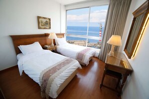 A hotel room with two beds, a nightstand, a desk, and a large window offering a view of the sea and a lighthouse. - Holiday Home Suites (Beirut)