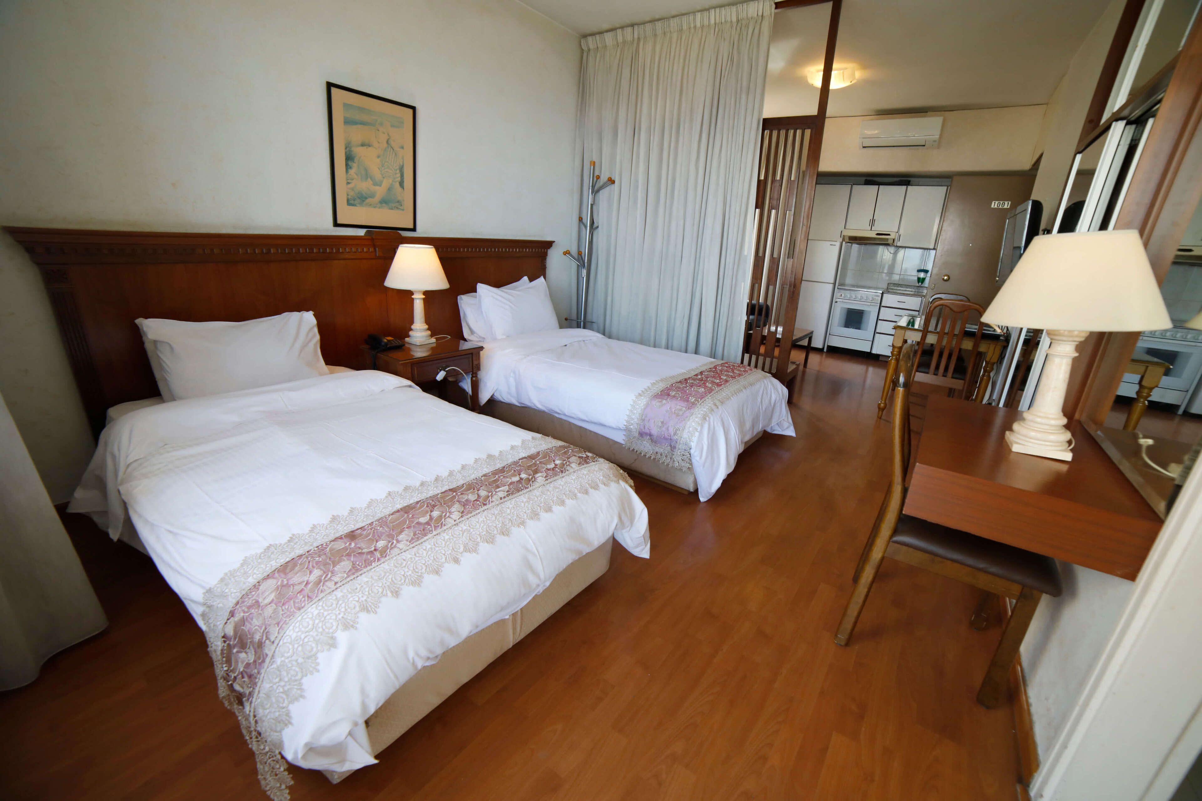 Habitación de hotel con dos camas, cabecera de madera, mesa de comedor con sillas, zona de cocina y un ventanal con cortinas.