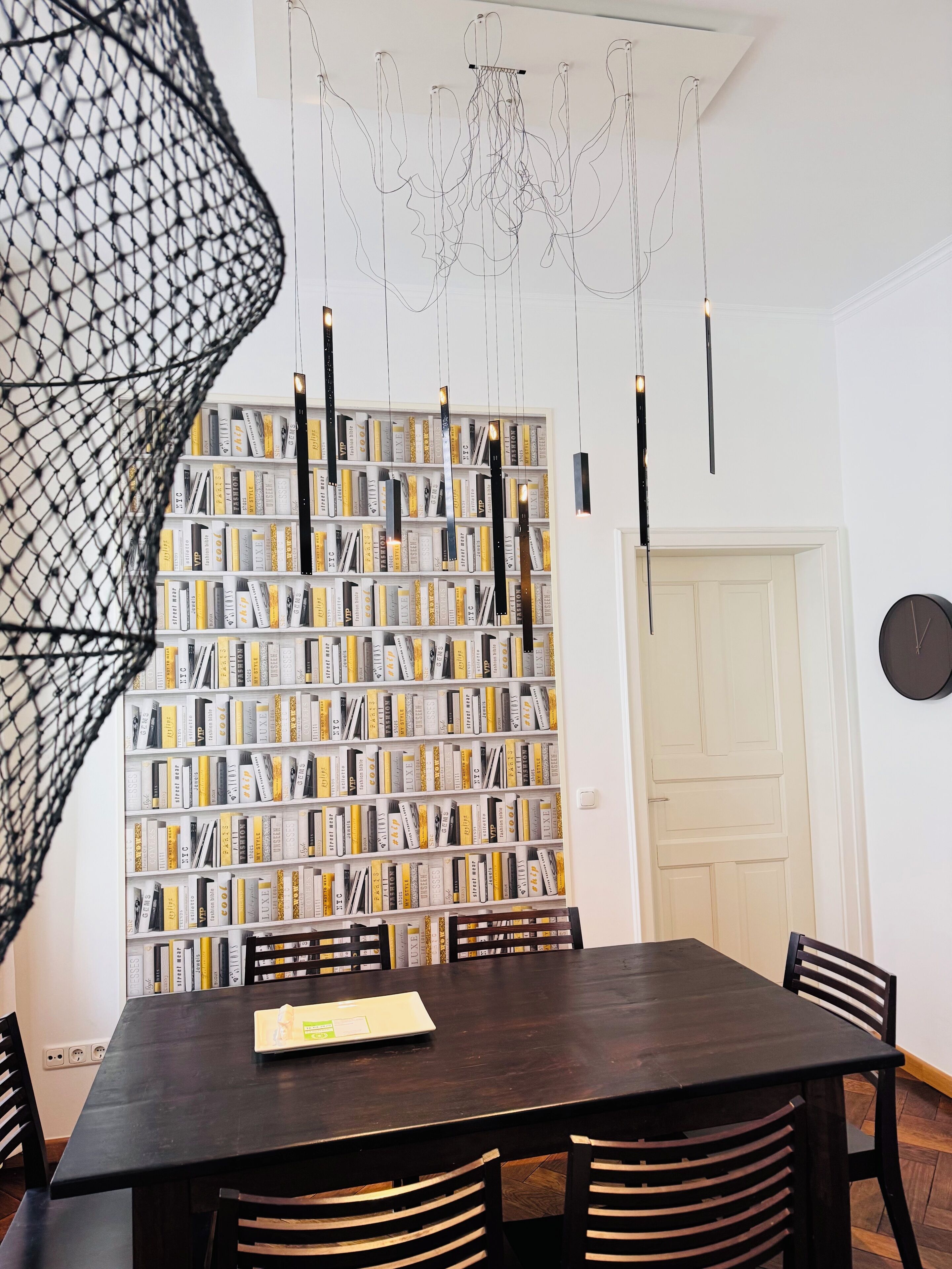 A dining area with a dark wooden table and chairs, a bookshelf filled with books, and a chandelier with multiple light fixtures.