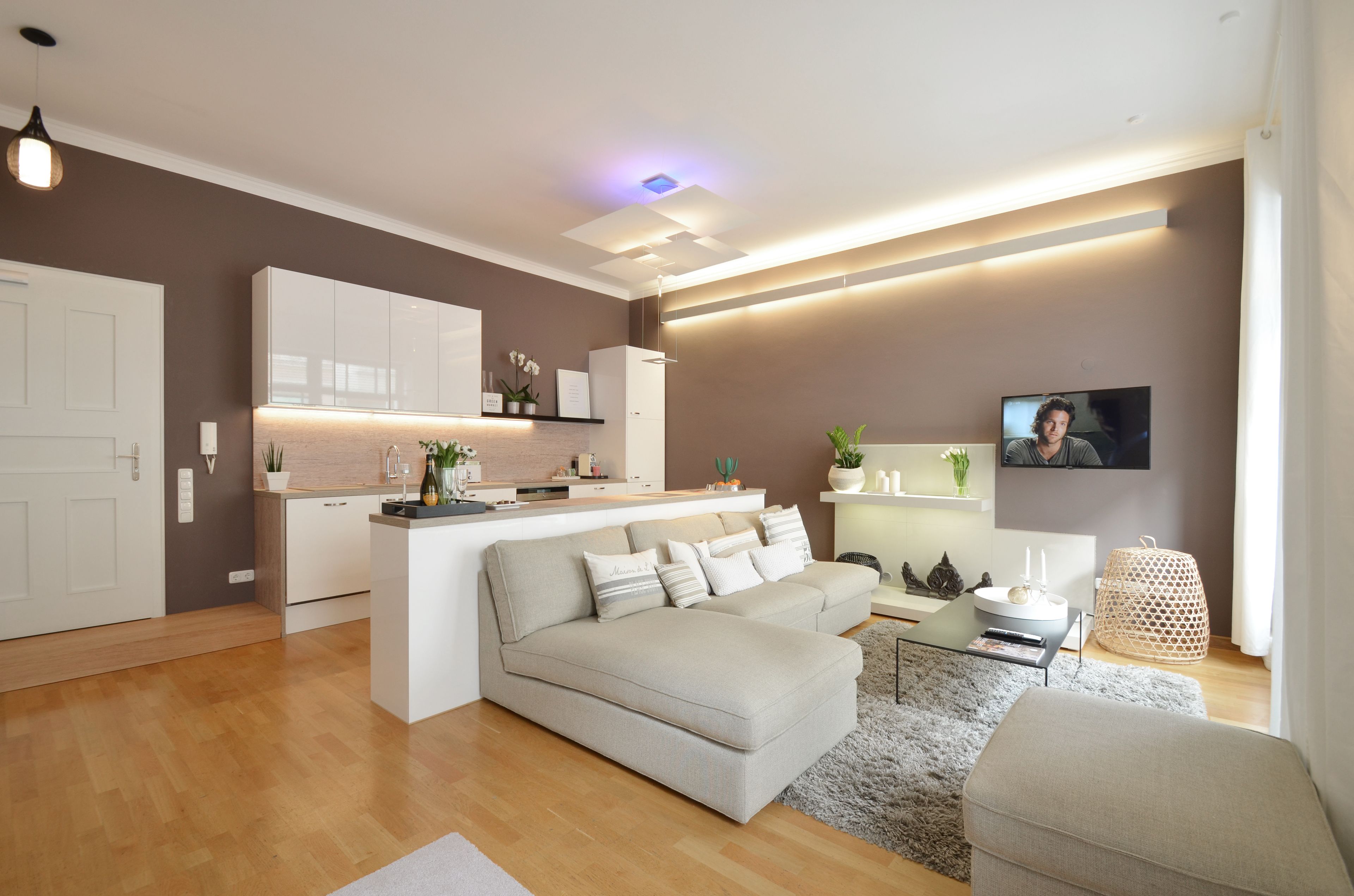 Una sala de estar moderna con un sofá, una mesa de centro y un televisor. Hay una zona de cocina con armarios y una encimera.