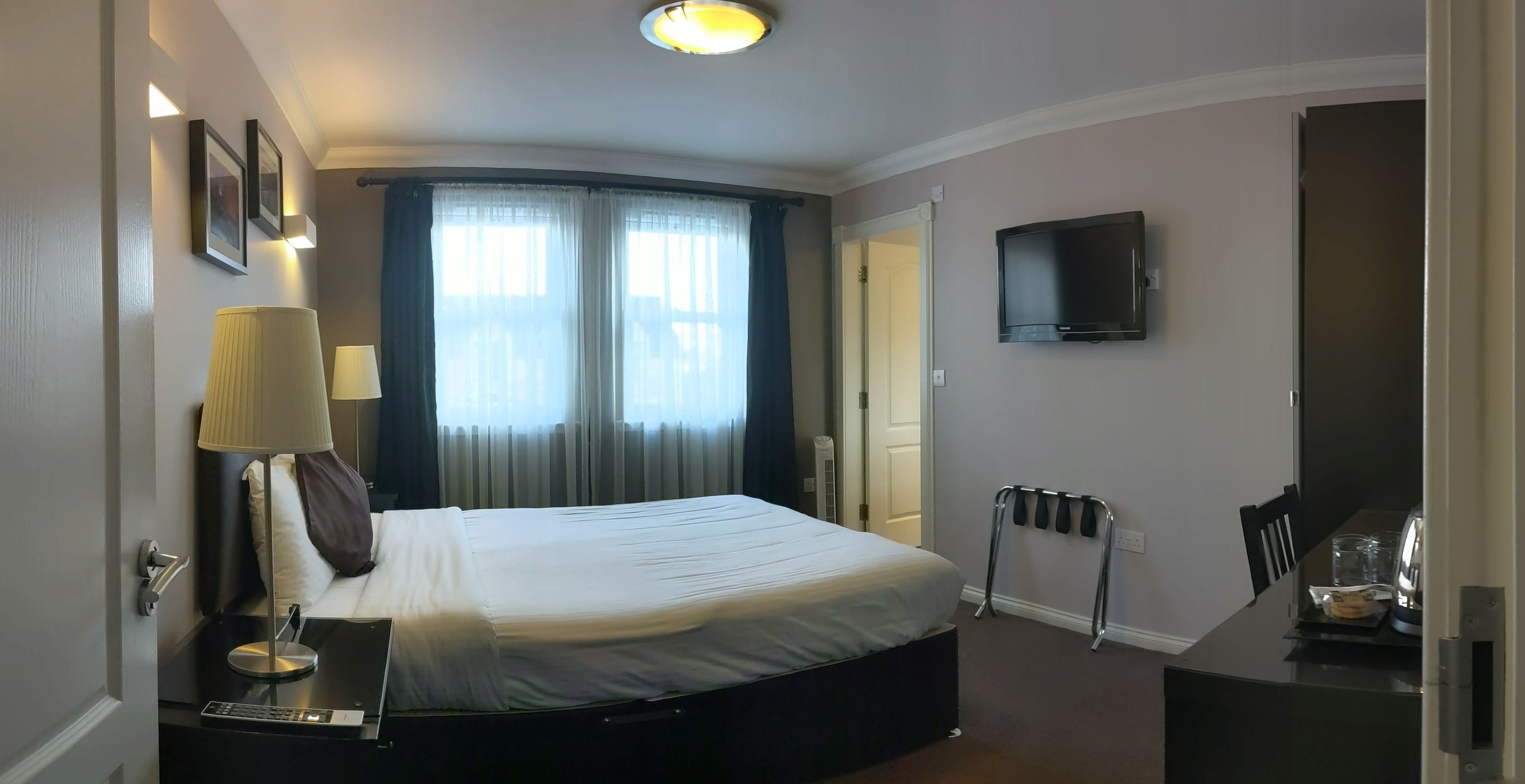 A hotel room with a bed, a desk with a lamp, a TV, and a chair.