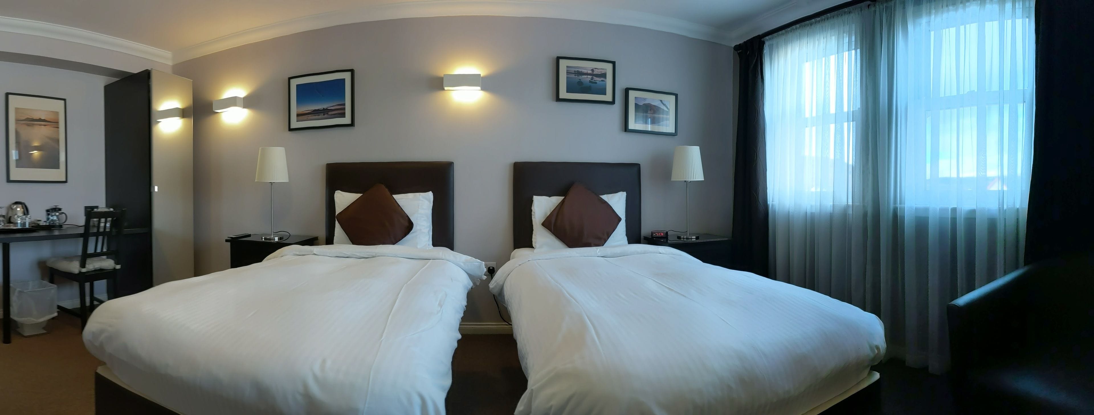 A hotel room with two beds, a desk with a chair, a lamp, and framed pictures on the wall.